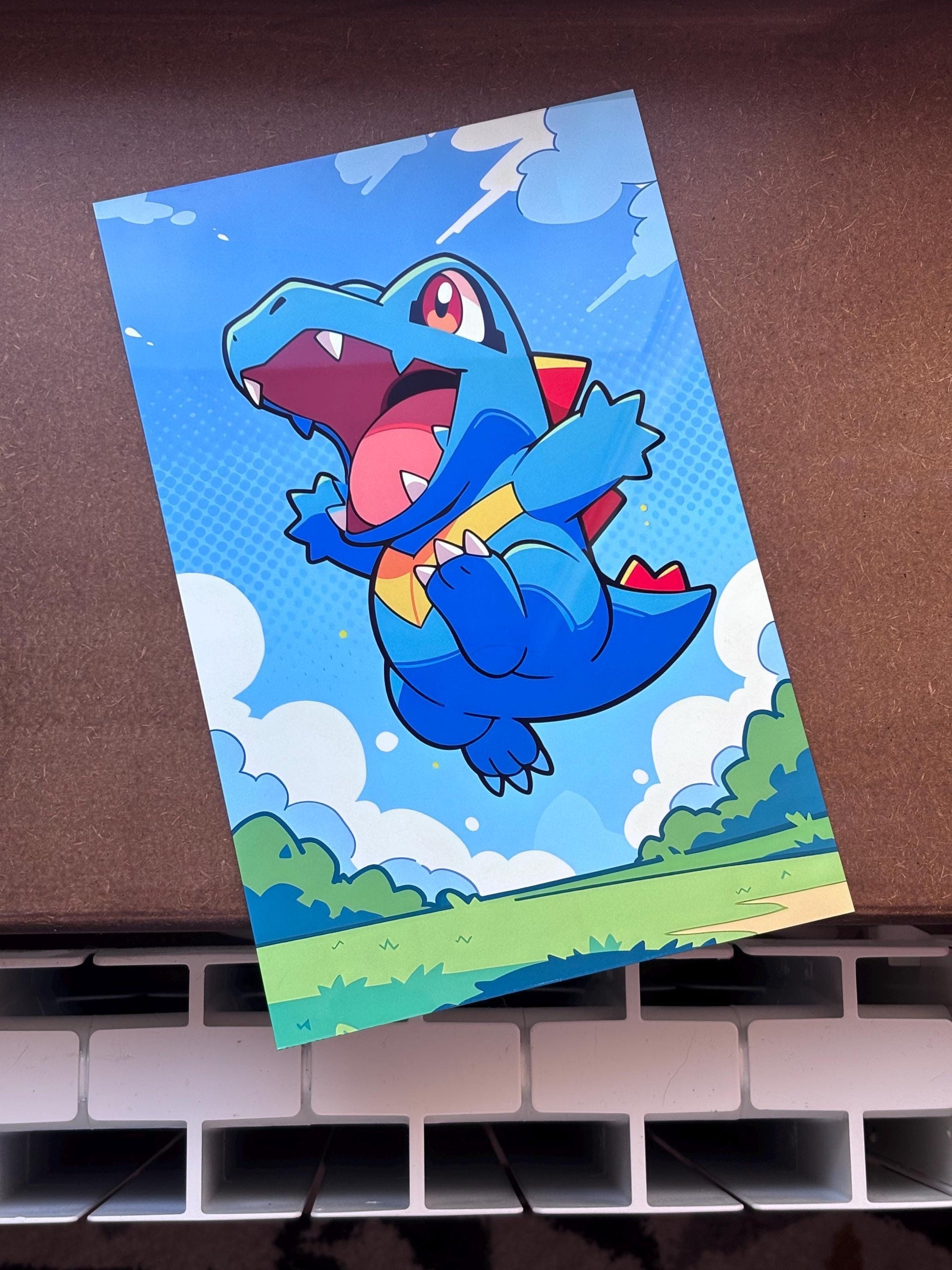 A colorful illustration of a cartoon-style crocodile character with a large open mouth, set against a blue sky with clouds.